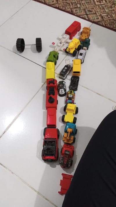 In Geulangganggampong, Indonesia, on a tiled floor, a collection of toy vehicles is arranged in a line and a separate pair of wheels. The main arrangement is a chain of toy cars, trucks, and construction vehicles. Starting from the top, a red truck-like vehicle is followed by a white airplane with red accents, a yellow cement mixer, a green truck, and a yellow truck. Below these, a black police car and a yellow truck are positioned. Further down, a red truck with a black interior is followed by a black car. Next is a colorful toy car with the number 36 on it, then a yellow construction vehicle, another yellow truck, and a blue construction vehicle. The line concludes with a red truck with a ladder, a red open-top vehicle, and a separate red toy piece on the floor. A single pair of black toy wheels connected by an axle is situated to the left of the main arrangement. The floor is white tile with visible grout lines, and a portion of a patterned rug is visible in the upper right corner of the image.