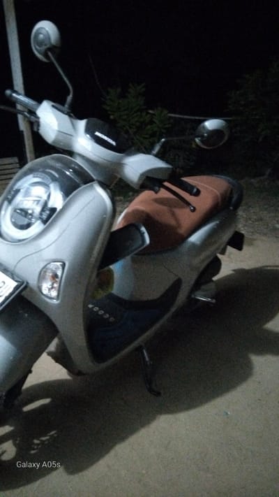 A silver Honda scooter is parked outdoors at night in Rambongpayong, Indonesia. The scooter is facing left and is partially illuminated by what appears to be street lighting or ambient light. The scooter has a brown seat and a prominent headlight assembly. The background is dark, with the shapes of trees and foliage visible, indicating an outdoor environment. The ground beneath the scooter is a dirt or gravel surface. A shadow of the scooter is cast on the ground. The "Galaxy A05s" watermark is visible in the bottom left corner of the image.