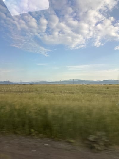 The image depicts a wide, open landscape under a sky filled with scattered clouds. In the foreground, a dirt road runs horizontally across the bottom of the frame. Beyond the road is a vast, grassy field, appearing somewhat blurred due to motion, suggesting the photograph was taken from a moving vehicle. The field stretches towards a distant horizon. In the mid-ground, several tall power transmission towers are visible, with power lines stretching between them. Behind these towers, a range of low, hazy mountains can be seen. The sky is a pale blue with white and light gray clouds. The overall scene is natural and rural, possibly a steppe or plains environment, and the presence of power lines suggests proximity to some form of infrastructure or development within a sparsely populated area. The light suggests it is daytime, likely late morning or afternoon. The image is taken from a slightly elevated perspective. There are no people or animals visible in the frame.