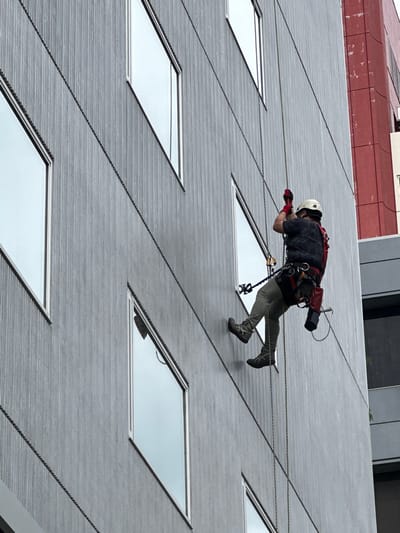 A single individual, an adult male, is depicted suspended vertically against the grey, vertically paneled facade of a multi-story building in Chūō, Japan. The person wears a light-colored hard hat, a dark patterned t-shirt, light khaki-colored trousers, and dark athletic shoes with light soles. Red gloves cover his hands, which are elevated, manipulating grey ropes that run diagonally down the building face. He is secured by a red and black full-body harness, from which a black cylindrical object (possibly a lens or tool) hangs on his right hip, and various rappelling/work-at-height devices are attached. A red backpack or equipment bag is visible on his back. His body is slightly angled, with his feet maintaining contact with the building's surface. The building features multiple rectangular windows with light frames, irregularly spaced. To the right, a section of an adjacent red building with a textured, weathered surface is visible, alongside another building with dark windows below it. The sky, reflected in the windows, appears overcast.