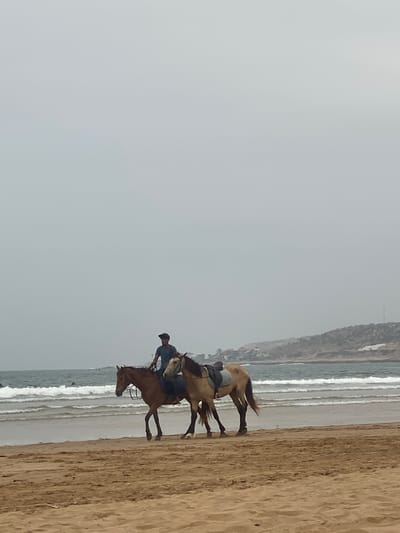 Two people are riding horses on the beach in Taghazout, Morocco. The sky is overcast. A ridge of land can be seen beyond the surf in the background.