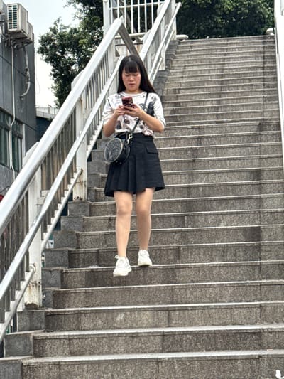 A young woman with long black hair stands on a staircase in 龙溪街道, China, holding a mobile phone and looking at the screen. She wears a patterned white t-shirt, a dark pleated skirt, and white sneakers. A small, round purse hangs from her shoulder. The staircase has a metal railing. A building with an air conditioning unit is visible in the background, along with trees.