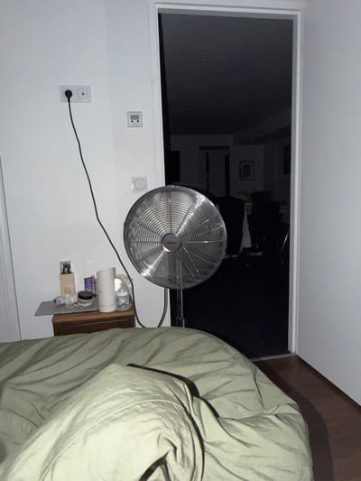 The image depicts an indoor scene, likely a bedroom, viewed from a low angle. In the foreground, a neatly made bed is covered with a light green comforter. To the left of the bed, a white wall features an electrical outlet with a black plug inserted into it, connected to a black cord that trails downwards. A small digital thermostat is mounted on the wall above a bedside table.
On the bedside table, several objects are arranged: a small figurine in a box, a cream-colored bottle, a white cylindrical object, and a baby bottle with a clear liquid. To the right of the bedside table stands a silver oscillating fan with a metallic grille, its power cord not visible.
The background reveals an open doorway leading into a dark room, where furniture and dark drapes are dimly visible, suggesting it could be a living room or another bedroom. The lighting in the scene is dim, contributing to the overall somber atmosphere. The environment suggests a residential setting.
There is no visible indication of the location being specifically in Pontonx-sur-l'Adour, France.