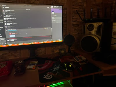 The image displays a dimly lit computer desk setup in a room with a brick-patterned wall. A prominent computer monitor shows a Discord chat interface. The chat, mostly in Bulgarian, features conversations with a user named "Dancho" with timestamps from late 2025 and early 2026. Notable messages include a Discord invite link and system notifications stating "Невалидна покана" (Invalid invite) and "Попитай Danchoо за нова покана!" (Ask Dancho for a new invite!). A Discord user profile for "Danche," registered on "18.07.2016," is also visible, showing "Общи сървъри - 2" (Shared servers) and "Общи приятели - 2" (Shared friends).
On the desk, a black gaming mouse with yellow LED accents is placed on a mouse pad featuring a graphic design and a "SteelSeries" logo. A pop filter on a stand is positioned in front of the monitor, suggesting microphone use. A mechanical keyboard with green backlighting is visible beneath the desk. To the left, an ashtray filled with cigarette butts and a cigarette pack are visible next to a red item. Headphones are resting on the right side of the desk.
To the right of the monitor, a black router with multiple antennas and green indicator lights is perched on top of a large, silver-grey speaker. Part of a computer tower with "XDSS" visible is next to the speaker. The dim lighting suggests it is evening or night. This setup is located in Vlado Trichkov, Bulgaria.