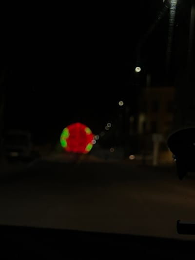 The image shows a blurred red spherical object with green markings hanging in a dark environment. It is unclear where it is located in the city of Sidi Kacem, Morocco. The object appears to be illuminated, standing out against the dark background. Other objects and shapes are visible in the background, but they are out of focus and difficult to identify.