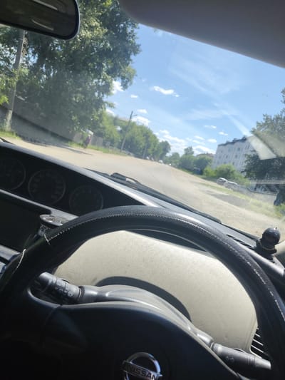 The image shows the interior of a car, looking out through the windshield at a street scene in the city of Shimanovsk, Russia. The road ahead is paved and appears to be in a residential area, with trees lining both sides. Several buildings are visible in the background, including a multi-story apartment building. The sky is blue with scattered clouds. The car's dashboard and steering wheel are in the foreground. The steering wheel has a textured grip and the Nissan logo is visible on it. A windshield wiper control stalk is also visible on the steering column. A small, decorative figurine of a person wearing a helmet is attached to the dashboard near the windshield. There is a person walking on the sidewalk in the distance. The overall impression is a snapshot taken while driving on a sunny day.