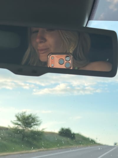 The image is a selfie taken in a car's rearview mirror. The primary subject is a young woman, reflected in the mirror, with blonde hair and her face tilted downwards. She is wearing a light-colored top. She is holding a rose-gold colored smartphone, which is also reflected in the mirror, displaying a triple-lens camera system.
The setting appears to be inside a car, looking forward through the windshield. The reflection in the mirror shows the woman and her phone. Outside the car, visible through the windshield and in the background reflection, is a bright sky with scattered clouds, suggesting it is daytime. The light-colored road and green trees on the side indicate a rural or suburban setting, possibly on a highway or a country road. The presence of some water droplets on the windshield suggests recent rain or high humidity.
The woman's expression is neutral, perhaps contemplative or relaxed. The overall mood is casual and personal, typical of a spontaneous selfie. The image's orientation is vertical, with the top of the frame showing the sky and the bottom showing the interior of the car and the reflection.
No visible text is present in the image. The location context of Utrennyaya Dolina, Russia, is not directly discernible from the visual elements alone but is provided as external information.