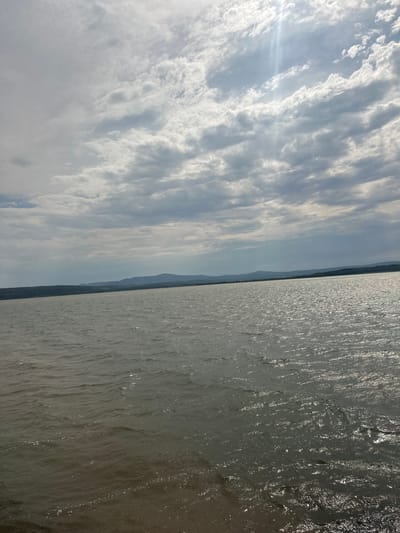 This image captures a landscape scene likely taken from a boat or waterfront. The composition is split horizontally, with the left side dominated by a vast sky filled with textured, wispy clouds. These clouds are illuminated by the sun, which creates a soft glow and lens flare in the upper left quadrant. The right side of the image features a body of water with a rippled surface, reflecting the sunlight in shimmering patterns. In the distance, a dark, mountainous horizon line separates the sky and water, suggesting a coastal or lakeside setting. The muted colors of the sky and the sunlit water create a serene and expansive atmosphere.
The image appears to be taken during the daytime, given the bright sunlight and visible cloud formations. The weather seems fair with some cloud cover, but not overcast. The landscape suggests a natural or semi-natural environment, potentially near a large body of water like a sea, lake, or wide river. The presence of a distant landmass implies a connection to land beyond the immediate foreground. No people, specific objects, or text are visible in the image. The overall mood is peaceful and reflective, emphasizing the grandeur of the natural elements.