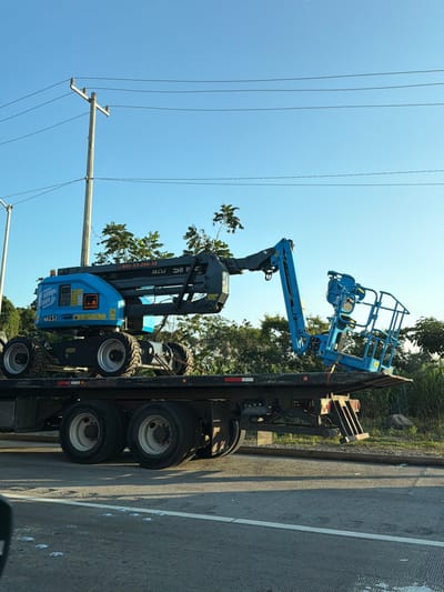A blue Genie AR15J boom lift sits secured on a black flatbed trailer, with its articulated arm and platform extended. The trailer is on a gray asphalt road, with a solid white line to the right. Utility poles with power lines run parallel to the road against a clear blue sky. Green trees are visible in the background. The lift and trailer appear to be stationary.