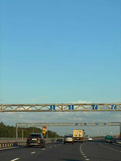The image depicts a highway scene in Zimnyaya Gorka, Russia, under a clear blue sky. In the foreground, several vehicles are traveling on the asphalt road. A dark-colored Lexus SUV is in the left lane, followed by a silver car in the middle lane and a white truck in the right lane. Further ahead, a green truck is visible in the right lane. Above the highway, overhead gantries display various road signs with directional arrows, indicating lane usage. On a lower gantry, a triangular warning sign with a silhouette of a person working and a circular sign with "70" are visible, suggesting a speed limit or construction zone. Trees line the right side of the highway, and a metal guardrail separates the lanes. The overall environment appears to be a well-maintained, multi-lane highway during daylight hours.