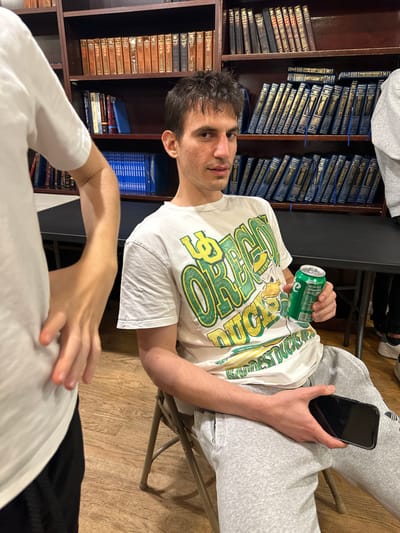 The image depicts an indoor scene in Bergenfield, United States, primarily focused on a male subject seated on a light-colored metal folding chair. The subject, appearing to be in his 20s or early 30s, has short, dark, disheveled hair, a slight mustache, and light facial stubble. He wears a white t-shirt with a green and yellow "Oregon Ducks" logo and light grey sweatpants. His right hand holds a green beverage can, consistent with Sprite, while his left hand rests on his lap, holding a black smartphone. He is looking directly towards the camera with a neutral expression.
The background consists of dark wooden bookshelves filled with numerous books. Many books feature uniform dark blue covers with gold lettering, suggesting a collection of religious texts or a specialized library. Other books with brown/tan bindings are also present. A dark table surface is visible behind the seated subject. Partially visible on the left is the torso and arm of another individual wearing a white t-shirt. On the far right, a third individual is partially discernible, wearing a grey hoodie and dark pants. The flooring is a light-toned wood laminate.