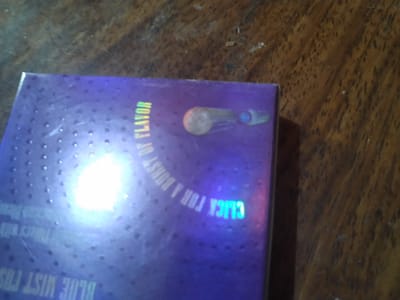 A close-up shot shows a purple box with a metallic sheen resting on a wooden surface. The box is adorned with a pattern of small, raised dots. A bright, white light source reflects off the surface of the box, obscuring some of the details. Text on the box is partially visible, including the words "FLAVOR" and "CLICK FOR A BURST." To the right of the text, a graphic depicting a light brown oval shape connected to a blue circular shape is present. The wooden surface appears to be a table or countertop with visible grain patterns and some specks of dust. The overall lighting is dim, with the bright reflection being the dominant light source in the immediate vicinity of the box. The image is an indoor scene, and no information about the city of Gampongbaro, Indonesia, is present.