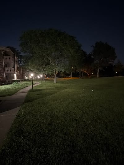 This is a nighttime, exterior photograph of a paved walkway curving through a grassy lawn. The walkway is illuminated by a series of street lamps, casting pools of light on the path and surrounding grass. To the left of the walkway, a multi-story building with balconies is visible. Several trees are scattered across the lawn, their foliage silhouetted against the dark sky. The scene appears to be within a residential or apartment complex in Vernon Hills, Illinois, United States of America.