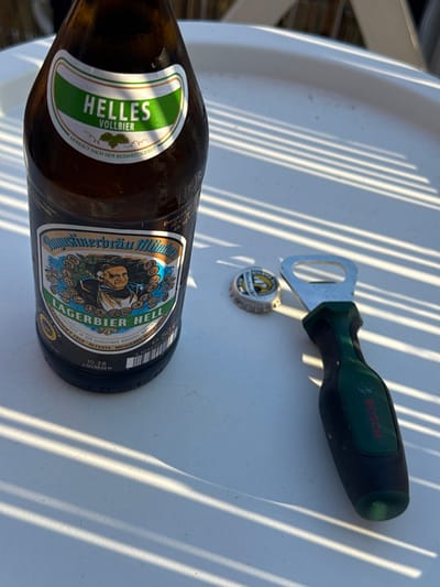 This media file shows a close-up, top-down view of a brown glass beer bottle and a Bosch bottle opener on a white, textured surface. The bottle is labeled "Helles Vollbier" and "Augustinerbräu München Lagerbier Hell," indicating it is a German lager beer. The bottle opener is green and black with the Bosch logo visible. A bottle cap, also with a beer logo, lies between the opener and the bottle. The scene is illuminated by bright sunlight, casting strong, parallel shadows across the surface, suggesting it is daytime. The surface appears to be outdoors, possibly a table. The overall impression is one of relaxation and enjoying a beverage, likely during warm, sunny weather. The prominent shadows and clear labels are notable details.