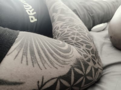 A close-up, monochromatic image shows a person's tattooed arm resting on a bed. The tattoo features geometric designs, including patterns reminiscent of flowers and grids. A portion of a black t-shirt with the word "PRO" visible is on the left side of the frame. A white object resembling a pillow or cushion is visible near the person's arm. The scene is indoors, presumably in a bedroom. There is no specific evidence to suggest that the image was taken in Lomas de Zamora, Argentina.
