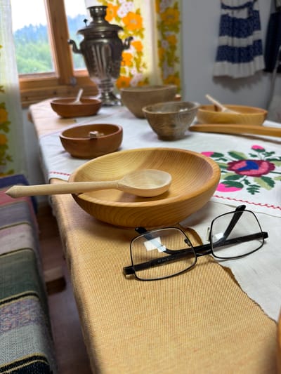 An indoor scene depicts a wooden table set with several handcrafted wooden bowls and spoons, a traditional Russian samovar, and a pair of eyeglasses. The table is covered with a tablecloth featuring embroidered flowers. The background reveals a window with a view of a green, mountainous landscape. The overall impression is one of a rustic, home setting, likely in a rural area, possibly a guesthouse or a domestic space in Ust'-Kabyrza, Russia. The lighting is natural, coming from the window.