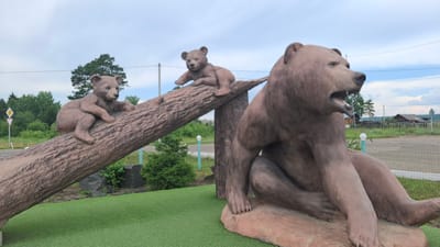 The image shows a sculpture of three bears in the village of Polovino-Cheremkhovo, Russia. The main subject is a large bear sculpture sitting on a rock, with its mouth open as if roaring. To the left, a log sculpture is positioned diagonally, with two smaller bear sculptures perched on it. One smaller bear is sitting near the top of the log, and another is positioned further down. The sculptures are situated on a green artificial turf surface. In the background, there is a residential area with wooden houses and a fence, surrounded by trees and a cloudy sky. A road and a white traffic sign are also visible.