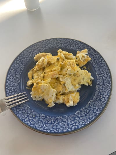A plate of scrambled eggs is placed on a white table. The plate is dark blue with a white patterned rim. A fork with four tines rests on the left side of the plate, partially submerged in the eggs. The eggs are fluffy and golden yellow, with visible herbs mixed in. The background shows a white surface with a soft shadow cast by an object out of frame to the top left. The lighting suggests an indoor setting, possibly a kitchen or dining area. The context of Skala Avdiron, Greece, implies a meal served in a Greek locale.