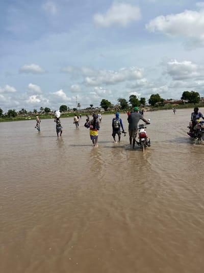 In Kunini, Nigeria, a group of people are wading through a flooded area. The water level reaches about knee-high for most individuals. Several people are carrying items on their heads, including what appears to be sacks and other bundled goods. Two individuals are riding motorcycles through the water, one in the foreground and another further back. Buildings and trees are visible in the background, suggesting a settlement or village. The sky is partly cloudy. The scene depicts a challenging environment due to the flooding, with people navigating the water with their belongings and some using motorcycles for transport.