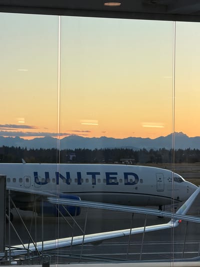 The image shows a United Airlines airplane parked at Sea-Tac International Airport in Seattle, Washington, USA. The plane's fuselage, with the "UNITED" logo clearly visible, is positioned in the foreground. Part of the wing and an engine are also visible. In the background, a scenic view of mountains and trees is seen against a golden-orange sunset sky, with clouds scattered across the horizon. A jet bridge extends towards the aircraft, and a portion of the airport terminal's glass facade is visible, reflecting the sky and the surrounding environment. The scene appears to be captured from inside the terminal, looking out at the tarmac.