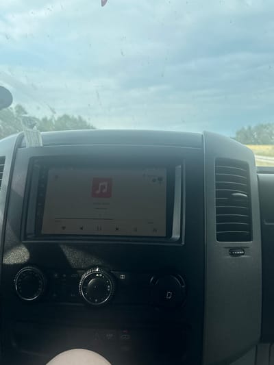The image shows the interior of a vehicle, likely a van or truck, facing forward. The central dashboard features a touchscreen display showing a music player interface with a musical note icon, song title "Костры Любви" (Bonfires of Love), and artist "Михаил Круг" (Mikhail Krug). Below the screen are climate control knobs and buttons, including "ASR OFF". The view through the windshield reveals a hazy, overcast sky and blurred trees in the distance, suggesting the vehicle is in motion. The overall scene is a close-up of the car's dashboard and windshield. The location is stated to be Konopki-Monety, Poland.