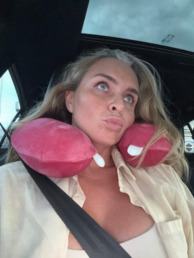 In the city of Ban Nong Mu, Thailand, a woman with blonde hair, gray streaks, and blue eyes is seated in a car. She is wearing a cream-colored collared shirt with the top buttons undone, revealing a white strapless top underneath, and a black seatbelt. Her head is supported by a pink neck pillow, and she is looking upwards with her lips pursed. The car's sunroof is visible, as is the interior of the vehicle.