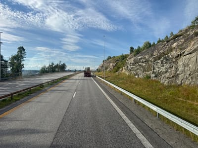 The image shows a scenic view of a highway in Espa, Norway. The highway is paved with asphalt and has multiple lanes, separated by white and yellow lines. A guardrail runs along the right side of the road, and a rocky, tree-covered hillside rises to the right. The sky is blue with scattered white clouds, suggesting it is daytime with fair weather.
In the distance, a red truck with a flatbed trailer is visible driving on the highway. There are no people clearly visible in the image. The overall setting is a highway cutting through a natural landscape, with the rocky hillside and trees indicating a rural or semi-rural location. The time of day appears to be during daylight hours, possibly late morning or afternoon, given the lighting and shadows. The clear sky and bright sunlight suggest pleasant weather conditions. There is no visible text in the image.