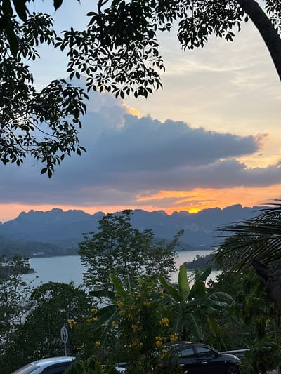 The image captures a sunset view from what appears to be the city of Ban Chiao Lan, Thailand, framed by lush greenery. The sky is a gradient of colors, transitioning from blue to orange and yellow as the sun dips behind the silhouette of distant mountains. There are large clouds filling a portion of the sky, with the sun's light piercing through a gap between the mountains. Foreground elements include tree branches and foliage, some with distinct yellow flowers. A glimpse of a body of water can be seen between the vegetation and the mountain range. Part of a vehicle is visible on the right side of the frame, partially obscured by the surrounding plants. A street sign is also visible by the car.