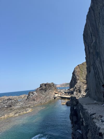 The image captures a sunny day with clear blue skies over a rocky coastline. In the foreground, large, dark gray rocks form an overhang, creating a sense of depth and framing the scene. Below this overhang, a section of the rocky coast descends towards the sea. The water is a vibrant turquoise near the shore, transitioning to a deep blue further out, with some visible waves breaking.

Further along the coastline, more rugged, dark-colored rocks are visible, some with patches of greenery on top. In the distance, a town with buildings can be seen nestled against a hillside. Several boats are present in the water near the town.

On the rocky shore in the middle ground, a few people are scattered, some appearing to be walking or sitting. The overall impression is one of a serene coastal landscape, likely during the daytime given the bright sunlight. The location appears to be Argelès-sur-Mer, France, as indicated by the context. No visible text is present in the image.