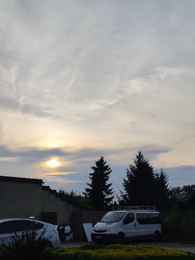 The image depicts an outdoor scene at dusk in Czapury, Poland. The sky is filled with a mix of clouds, with the sun partially visible and casting a warm glow. The setting appears to be a residential or semi-rural area, with a building on the left and trees silhouetted against the sky.
In the foreground, several objects are visible. A white van with a roof rack is parked on the right, and a white car, possibly a hybrid, is parked on the left. The car has the number "14145" visible on its side, along with a checkered pattern, suggesting it might be a taxi or a service vehicle. There are also some clothes hanging to dry near the building on the left.
The overall mood of the image is calm and serene, characteristic of the end of the day. The lighting suggests it is twilight or early evening. The presence of the building, vehicles, and laundry indicates a domestic or everyday setting. The silhouetted trees add a natural element to the scene, contrasting with the man-made objects. There are no people clearly visible in the image.