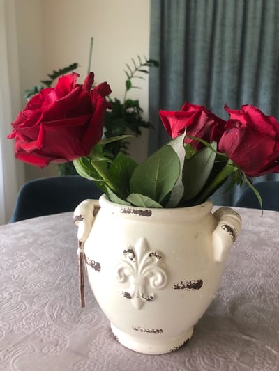 The image displays a close-up, vertically oriented view of a bouquet of red roses in a cream-colored, distressed ceramic vase. The vase has ornate detailing, including what appears to be a fleur-de-lis emblem and textured patches that mimic rust or age. The roses themselves are a deep red, with some petals showing water droplets. Green leaves are interspersed among the blooms.
The vase is placed on a textured, light-colored tablecloth with a subtle floral pattern. In the background, a portion of a teal curtain with horizontal pleats is visible, and to the right, a sheer white lace curtain can be seen. A dark chair is partially visible in the lower right quadrant of the image. A thin, twig-like object is positioned horizontally across the front of the vase.
The lighting suggests it could be daytime, possibly indoors with natural light. There are no people or animals present. The overall mood is one of still life, focusing on the beauty of the flowers and the decorative vase. There is no visible text in the image. The location context provided is Šeškai, Lithuania.