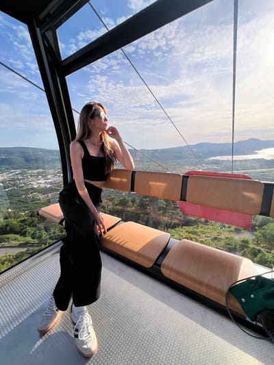 The image displays a lone female subject inside a modern cable car cabin, observing the panoramic landscape of Čavori, Montenegro. She has long, light brown hair and wears green-framed sunglasses, a black square-neck crop top, black wide-leg pants, and light-colored Adidas sneakers with white socks. She is seated on a light brown wooden bench, leaning against its backrest, with her left hand resting on her chin as she looks out towards the right side of the frame. The cable car interior features large glass windows, black structural framing, and a textured metallic floor. Through the windows, a verdant, hilly landscape extends below, dotted with a town characterized by numerous buildings. In the distance to the right, a body of water reflects sunlight under a blue sky with scattered white clouds. A green object is partially visible on the far right bench.