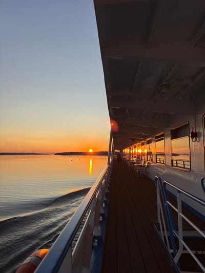 The image depicts a view from the deck of a ship sailing on a body of water at sunset. The sun, a bright orange orb, is low on the horizon, casting a warm glow across the sky and water. The sky transitions from a pale blue at the top to a vibrant orange near the horizon. Silhouetted against the sky are distant landforms, appearing as dark, flat shapes.
The right side of the image shows the side of the ship, with a long row of windows reflecting the sunset. Tables and chairs are visible on the deck in front of the windows. A walkway with railings runs along the side of the ship. The deck itself is wooden.
In the water, a wake trails behind the ship, creating ripples and patterns on the surface. A life preserver is visible near the railing in the foreground.
There is one person visible walking along the deck in the distance. The scene appears to be serene and peaceful. There is no information provided within the image itself to indicate a specific location within the city of Шайма, Russia.