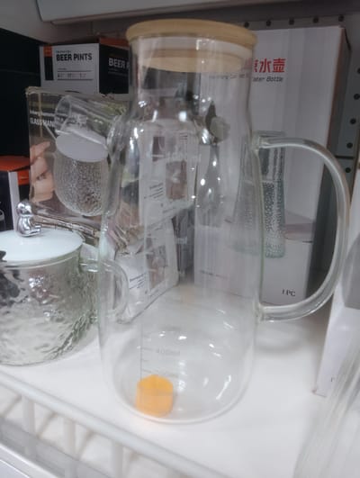 The image displays a close-up view of a glass pitcher with a wooden lid, placed on a white shelf. The pitcher has measurement markings on its side, indicating volumes such as 200ml, 400ml, and 500ml. A small orange cap is visible inside the pitcher near the bottom. Behind the pitcher, boxes with text and images are stacked. One box has "BEER PINTS" written on it, and another has Chinese characters and the words "ater Bottle". To the left of the pitcher, a textured glass jar with a lid and a silver decorative knob is partially visible. The overall scene appears to be a retail environment, likely a shelf in a store. The specific location in the city of Aketu-Oja, Nigeria, is not deducible from the visual information provided in the image.