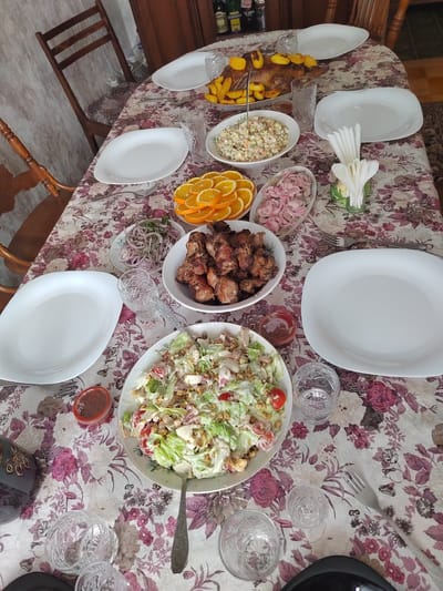 A dining table covered with a floral patterned tablecloth is laden with food. Several white serving dishes hold various items including a large salad, cooked meat pieces, sliced oranges, pickled onions, and another salad with mayonnaise. There is also a platter containing what appears to be roasted poultry and potatoes. A white plate, cutlery and various glasses are laid at multiple spots around the table. There are bottles of liquid towards the top edge. The table is surrounded by wooden chairs.
The scene appears to be set indoors and could be located in Италмас, Russia.