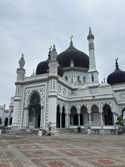 The image shows the Zahir Mosque in Alor Setar, Malaysia, under an overcast sky. The mosque is predominantly white with dark domes. It features intricate architectural details, including multiple minarets, arches, and decorative patterns. A paved courtyard with a geometric brick pattern is in the foreground. Benches and planters are visible in front of the mosque. No people are present in the image.