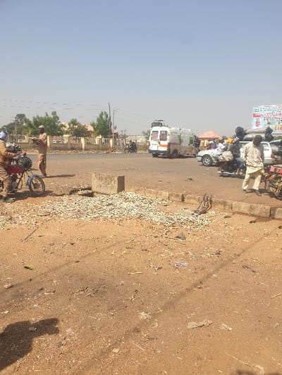 The image captures a sunlit, outdoor scene in Kafuri, Nigeria, depicting a busy, unpaved roadside area adjacent to a paved road under a clear, light blue sky.
The immediate foreground consists of dry, reddish-brown earth, extensively covered with light-colored, fragmented organic debris, possibly wood chips or plant matter, concentrated heavily towards the left-center. A rectangular concrete block rests on this ground in the left-center. A dark shadow, indicative of the photographer, is visible in the bottom-left corner.
In the left mid-ground, a red motorcycle is parked, with an individual wearing a blue helmet and brown attire seated on it, facing right. Another person in light brown attire stands immediately beside the motorcycle, also facing right. Further left, a third individual, similarly dressed in light brown, stands alone with arms slightly extended, facing right.
A paved road traverses the mid-ground horizontally, separated from the unpaved area by concrete curbs. On this road, a white van with green markings and a roof-mounted satellite dish or equipment rack, bearing the name "DANA," is positioned, facing right. Behind the van, a white sedan, visibly overloaded with items secured to its roof, is also present. Multiple individuals are visible around these vehicles; one person in a light-colored tunic and trousers stands near the white car, facing left. Various motorcycles, some with riders, are interspersed among the vehicles.
The background features several structures: a light-colored building wall with green trees behind it on the left, and other scattered buildings further right. A tall utility pole with overhead wires is prominent on the left side of the road. On the far right background, a billboard with partial text and imagery is visible. The scene is brightly lit by direct sunlight, and the ground appears dusty.