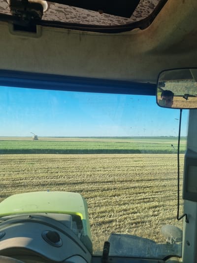The image depicts a view from inside a vehicle, likely a tractor or combine harvester, situated in a field near Lomnitsa, Bulgaria. The vehicle's interior is visible, including the dashboard, window, and mirror. Outside, a harvested field stretches into the distance, alongside a field of green plants. Another harvester is visible in the distance. The sky is clear and blue. The reflection of a person can be seen in the mirror.