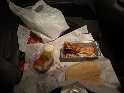 The scene depicts takeaway food arranged on a dark surface, consistent with the interior of a vehicle, in Hadleigh, Babergh, United Kingdom. The primary subject is a meal from "Babergh Kebab House" as identified by a visible receipt detailing the address "38 High St, Hadleigh, Ipswich IP7 5AL" and an order for "IAN NOBLE."
The food components include:
1. An aluminum foil container holding several pieces of cooked chicken, likely shish kebab, alongside a small portion of flatbread. Some chicken pieces exhibit charring.
2. A clear plastic container, with its hinged lid partially open, containing a salad composed of sliced tomatoes, cucumber, and one green pepperoncini.
3. A small, clear plastic cup filled with a white, creamy sauce.
4. A larger, irregularly shaped piece of flatbread (pitta) resting on white paper, with a visible bite taken from its edge.
The food items are placed on white greaseproof paper. To the left, a crumpled white plastic carrier bag is present, partially obscuring other items. A dark, upholstered car seat is visible on the far left. The overall lighting is dim. No individuals are present in the image.