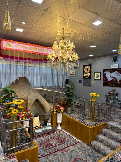 The image shows the interior of a Sikh Gurdwara, likely located in Chumkar, India. The focal point is a large, natural rock formation resembling a mountain, with an opening at its base. Surrounding this rock are various religious artifacts and decorative elements.
In the foreground, to the left, are several potted plants, including sunflowers and other colorful flowers. A metallic railing separates this area from a raised platform. The platform is carpeted and features a decorative border. To the right of the rock, there's a low silver structure, possibly a religious offering box or display, placed on the carpeted floor. A stringed musical instrument, similar to a tanpura or sitar, is propped against the rock.
The walls of the Gurdwara are adorned with religious artwork, maps, and framed texts. Notably, there's a large map depicting the journeys of Guru Nanak, the founder of Sikhism, and a framed Sikh Khanda symbol. Chandeliers made of gold and crystal hang from the ceiling, along with decorative tassels. The ceiling itself is a tiled surface with integrated square lighting panels.
The flooring throughout the Gurdwara is covered with intricately patterned Persian-style carpets in shades of red, blue, and grey. There are also steps with carpeted risers leading up to another level on the right side of the image. The overall ambiance is one of reverence and spiritual significance. No people are visible in the image.