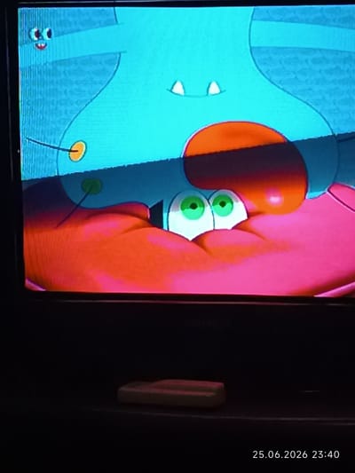 The image is a close-up, slightly distorted view of a television screen displaying an animated cartoon. The cartoon characters are predominantly turquoise and bright pink. Two large, circular green eyes with black pupils are prominent in the foreground, appearing to belong to a pink creature. Above these eyes, a large turquoise shape, possibly the body of another creature, is visible, with two small white triangular shapes resembling teeth or horns. A dark orange, oval-shaped area is positioned above the eyes. The background of the cartoon is a textured light blue. The television screen itself is in focus, showing static-like vertical lines. Below the screen, the dark surface of the television's casing is visible, with a small, rectangular object, possibly a remote control or a decorative item, placed on it. The timestamp "25.06.2026 23:40" is superimposed on the bottom right corner of the image, indicating a future date and time. There is no visible indication of the city of Bolhrad, Ukraine, within the image itself.