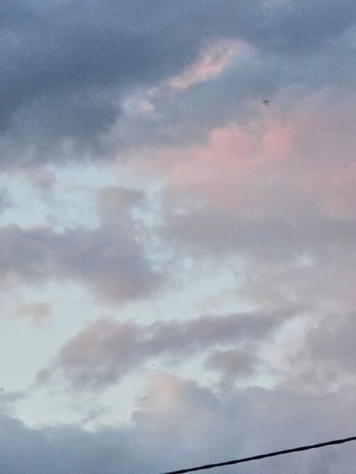 The media file shows a sky with clouds. The clouds are varied in color, with shades of pink, white, and grey, suggesting either sunrise or sunset. The overall lighting indicates that it is likely early morning or late evening. There is a thin, dark line visible on the right side of the frame, which could be a power line or a tree branch. The sky appears vast and open. The scene depicts a natural landscape, likely an outdoor setting in Satbayev, Kazakhstan. There are no people or explicit objects visible besides the potential power line. The mood is tranquil and serene, typical of a sky at dawn or dusk. There is no visible text in the image.