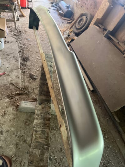 The image shows a light gray car bumper, possibly primed for painting, resting on a wooden support structure. The bumper is elongated with a gentle curve, and its surface is smooth and reflective. It appears to be positioned outdoors, in what looks like a workshop or garage setting, given the presence of wooden planks, dirt, debris on the ground, and some tools and equipment visible in the background.
In the background, a spare tire is visible, along with what appears to be an air compressor tank. There are also some wooden structures and sheets of material, suggesting a construction or repair environment. The lighting suggests it might be daytime, with natural light filtering into the space, though the area appears somewhat dim. The overall scene gives the impression of a work-in-progress, focusing on the preparation or repair of the car part. No people are clearly visible in the frame. There is no visible text in the image. The setting suggests a rustic or functional workspace, possibly in a rural area like Dragoevo, Bulgaria, given the context.