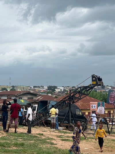 This image depicts an outdoor scene in Abeokuta, Nigeria, under an overcast sky suggesting a cloudy or possibly rainy day. A prominent black crane dominates the left side of the frame, its metal structure stretching upwards. Below and around the crane, several people are visible, appearing to be gathered.

In the foreground, a woman in a blue and gold patterned dress stands, looking towards the right. To her left, a group of people are clustered together, including a man in a red shirt, a person in a white shirt, and individuals in casual attire. Further to the right, a young boy in a yellow shirt and dark pants walks away from the camera, while another person in white stands near a banner.

Behind the people and the crane, a sprawling townscape with numerous buildings and rooftops stretches into the distance. The architecture suggests a densely populated urban area. Several large banners are visible, including one with a portrait of a man wearing a traditional cap and another with a portrait of a man in ceremonial attire. Text on one banner reads "Chief Kamarudeen".

The overall impression is of a public gathering or event, possibly related to a political campaign or local ceremony, given the banners and the presence of a crane, which might be used for staging or decoration. The mood appears casual, with people engaged in various activities within the gathering. The time of day seems to be daytime, but the cloud cover obscures direct sunlight.