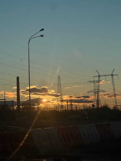 The image, correctly oriented horizontally, depicts an outdoor landscape scene during sunset or sunrise in Zelenodolsk, Russia. The sky occupies the upper two-thirds of the frame, transitioning from a pale blue at the top to warm orange and yellow hues near the horizon. The sun is prominently visible on the right side, low on the horizon, casting a bright glare and creating lens flares. Scattered clouds are illuminated from below, displaying warm orange and pink tones.
Several large electricity transmission pylons are silhouetted against the glowing sky, extending from the middle ground into the background. Numerous power lines stretch across the frame, connecting these pylons. In the left foreground, a modern street light with two curved arms and two lamps stands silhouetted against the bright sky. A dark, silhouetted line of trees or dense vegetation marks the distant horizon.
The lower part of the image is dark, suggesting ground level or a road. A faint white car can be discerned in the far distance on the right. On the extreme right foreground, a dark, vertical structure, possibly a barrier or part of a fence, is partially visible, showing hints of red and white stripes. No specific activity is taking place, and no visible text is present in the image.