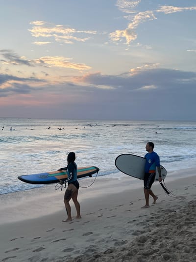 Two surfers, a woman and a man, are walking along the beach in Kuta Selatan, Indonesia, carrying their surfboards. The woman, on the left, wears a dark long-sleeved rash guard and black bikini bottoms. She has tattoos on her legs and is carrying a blue, orange, and yellow surfboard. The man, on the right, wears a blue and turquoise rash guard and patterned shorts. He is carrying a white surfboard. They are walking on a sandy beach with footprints visible. The ocean is to their left, with gentle waves breaking on the shore and several other surfers in the water. The sky above is filled with soft, pastel-colored clouds, suggesting it is either sunrise or sunset. The overall scene depicts a tranquil moment of surfers enjoying the ocean.