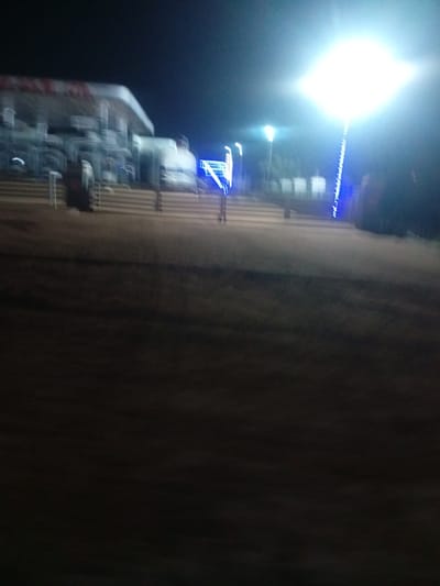 The media file captures a motion-blurred nighttime scene at what appears to be a gas station in Tundun Wada South, Nigeria. The primary subject is a gas station canopy with visible pumps and vehicles underneath, heavily blurred due to movement. To the right, a bright, diffused light source, likely a tall lamp post, dominates the sky, casting a blueish glow. Another smaller light source is visible to the left of the main lamp post, also appearing as a bright, indistinct light. The ground in front of the gas station is a mixture of light-colored paving and darker soil or sand. Blue string lights are intermittently visible along the base of the gas station structure and a pole. The overall impression is one of low light conditions and rapid movement, making specific details difficult to discern. The sky is dark, indicating it is nighttime. No people are clearly identifiable due to the motion blur. No legible text is visible in the image.