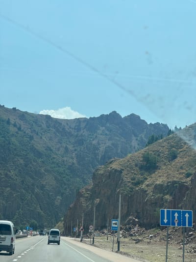 The image captures a scenic view of a highway passing through a mountainous region, possibly near Beshkul, Uzbekistan, given the provided context. The road is paved and appears to be a multi-lane highway with clear markings. Several vehicles are visible on the road, including white vans and cars, suggesting typical traffic flow.
The surrounding environment is dominated by rugged mountains with sparse vegetation, primarily dry grasses and shrubs. Rocky outcrops and steep slopes characterize the terrain. The sky is clear and blue, indicating fair weather.
Road signs are present along the highway, including directional signs indicating traffic flow and a speed limit sign showing "80". Another sign indicates "RADAR", likely a warning about speed enforcement. The presence of these signs and the well-maintained highway suggest a developed infrastructure within this natural landscape.
The image appears to be taken from inside a vehicle, likely from the perspective of a passenger or driver, as indicated by the slight distortion and the presence of the car interior elements. The overall impression is one of travel through a beautiful, albeit arid, mountainous area.