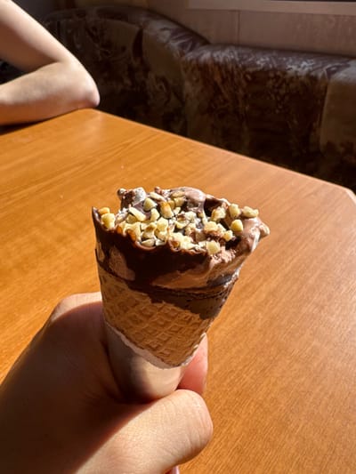 The scene is an interior view, located in Fowlis Easter, United Kingdom. In the foreground, a right hand holds a waffle ice cream cone. The ice cream is dark-colored, possibly chocolate, topped with numerous light-colored chopped nuts and a dark chocolate drizzle or shell. The top of the ice cream appears slightly irregular, suggesting a recent bite or partial melting. The cone is partially wrapped in white paper or foil at its base. The hand and cone cast distinct shadows to the right on a light brown wooden table, indicating strong illumination from the left. The table surface displays a visible wood grain. In the upper left quadrant, the bare forearm and elbow of a second individual are visible, resting on the same table. The background, largely out of focus, reveals a section of an upholstered sofa or bench with a dark, patterned fabric.