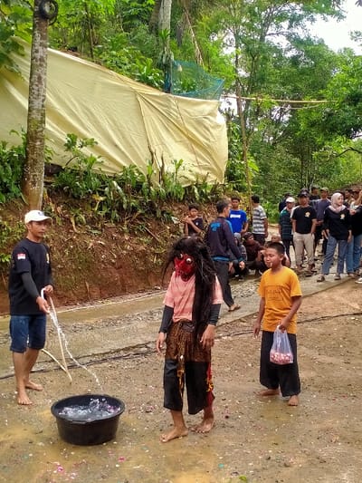 In Gempolan, Indonesia, an outdoor scene depicts a group of individuals engaged in an activity. In the foreground, a barefoot man wearing a white cap and dark t-shirt holds a white hose, directing a stream of water into a black plastic basin containing water and scattered pink/red objects. Adjacent to him, a barefoot figure in a red, grotesque mask with long, dark, matted hair stands, wearing a pink top, a brown patterned lower garment, and dark pants. This figure's lower right pant leg is discolored red. To the right, a barefoot boy in an orange t-shirt holds a clear plastic bag containing small red objects. The ground is wet, muddy dirt with some paved sections, and scattered with red/pink petals. In the midground, a crowd of approximately 15-20 individuals, including men, women (one wearing a hijab), and children, observe from behind the main figures. Behind them, a steep, heavily vegetated hillside features a large, light-yellow tarpaulin covering a structure, with some green netting visible above. Lush green trees and foliage form the background.